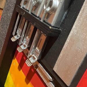 Może przedstawiać: Zbliżenie ręcznie robionej gitary basowej z tęczowym korpusem w paski. Instrument ma srebrny osprzęt i czarne akcenty. Widoczne są struny, a korpus pomalowany jest w czerwone, pomarańczowe, ż&oacute;łte i zielone paski. Gitara wydaje się dobrze wykonana.