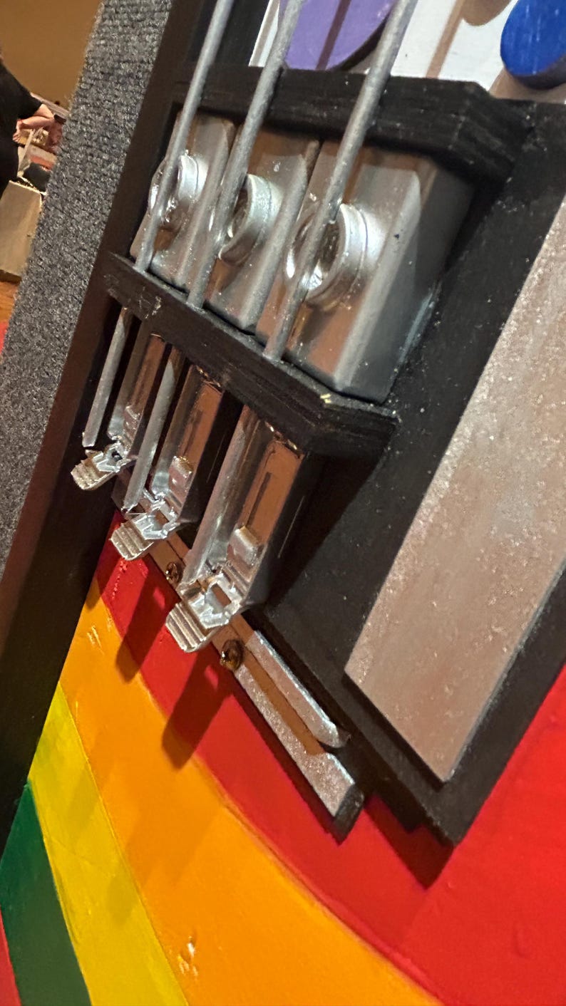 Może przedstawiać: Zbliżenie ręcznie robionej gitary basowej z tęczowym korpusem w paski. Instrument ma srebrny osprzęt i czarne akcenty. Widoczne są struny, a korpus pomalowany jest w czerwone, pomarańczowe, ż&oacute;łte i zielone paski. Gitara wydaje się dobrze wykonana.