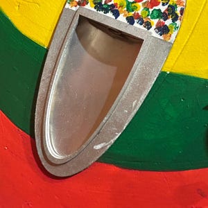 Może przedstawiać: Owalny, srebrny przedmiot z prostokątnym otworem, ozdobiony kolorowymi plamami farby. Obiekt umieszczony jest na tle czerwonych, zielonych, ż&oacute;łtych i pomarańczowych pas&oacute;w, tworząc żywą, artystyczną kompozycję.