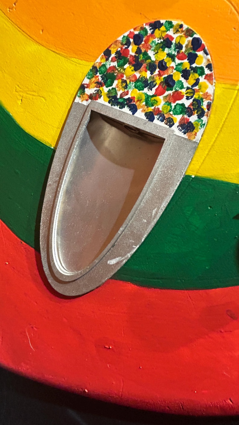 Może przedstawiać: Owalny, srebrny przedmiot z prostokątnym otworem, ozdobiony kolorowymi plamami farby. Obiekt umieszczony jest na tle czerwonych, zielonych, ż&oacute;łtych i pomarańczowych pas&oacute;w, tworząc żywą, artystyczną kompozycję.