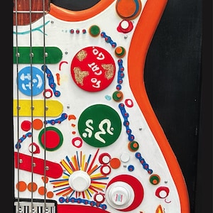 Może przedstawiać: Kolorowa, ręcznie malowana gitara basowa z tęczowym wzorem i r&oacute;żnymi elementami dekoracyjnymi. Gitara ma biały korpus z pomarańczowymi wykończeniami, czerwony pickguard i drewnianą szyjkę. Korpus zdobią koła, linie i napis "GO CAT GO."