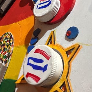 Może przedstawiać: Abstrakcyjne dzieło sztuki przedstawiające pomalowane zakrętki do butelek z czerwonymi, białymi i niebieskimi wzorami. Dzieło sztuki zawiera czerwone serca, ż&oacute;łte gwiazdy i tęczową sekcję, tworząc żywą i teksturowaną kompozycję.