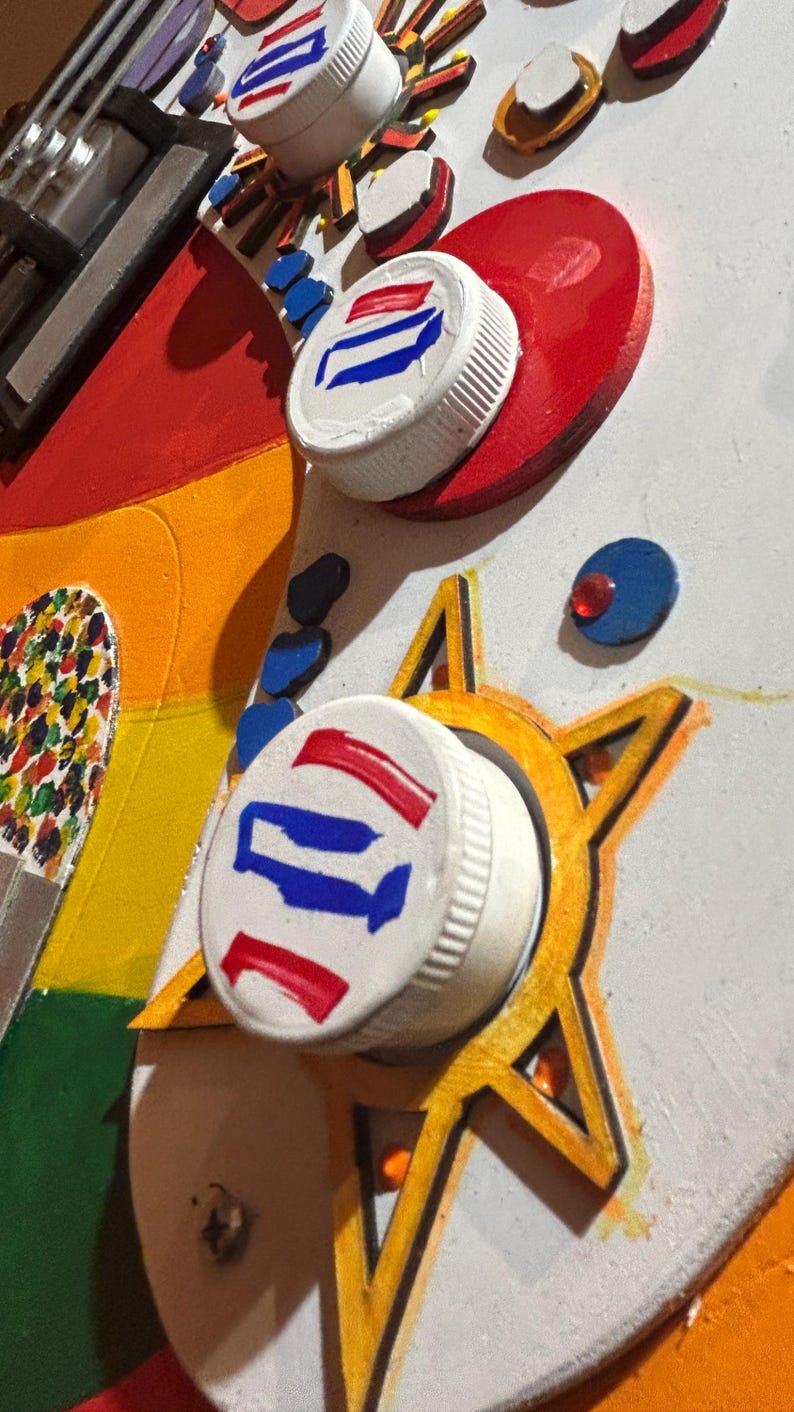 Może przedstawiać: Abstrakcyjne dzieło sztuki przedstawiające pomalowane zakrętki do butelek z czerwonymi, białymi i niebieskimi wzorami. Dzieło sztuki zawiera czerwone serca, ż&oacute;łte gwiazdy i tęczową sekcję, tworząc żywą i teksturowaną kompozycję.