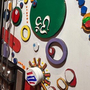 Może przedstawiać: Kolorowe, abstrakcyjne dzieło sztuki z motywem gitary. Dzieło zawiera r&oacute;żne kształty i kolory, w tym czerwony, ż&oacute;łty, zielony i niebieski. Zawiera białą zakrętkę z czerwonymi i niebieskimi paskami oraz zielone koło z białym tekstem.