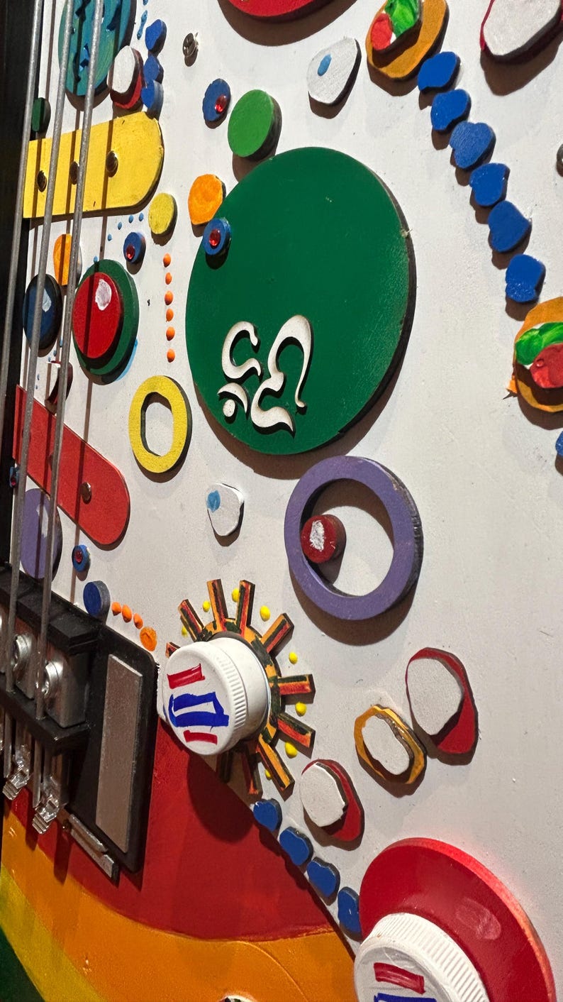 Może przedstawiać: Kolorowe, abstrakcyjne dzieło sztuki z motywem gitary. Dzieło zawiera r&oacute;żne kształty i kolory, w tym czerwony, ż&oacute;łty, zielony i niebieski. Zawiera białą zakrętkę z czerwonymi i niebieskimi paskami oraz zielone koło z białym tekstem.