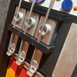 Może przedstawiać: Zbliżenie kolorowego, abstrakcyjnego dzieła sztuki przypominającego gitarę basową. Grafika przedstawia tęczowy korpus, srebrne struny i metalowy osprzęt. Elementy dekoracyjne obejmują fioletowe, niebieskie i czerwone koła.