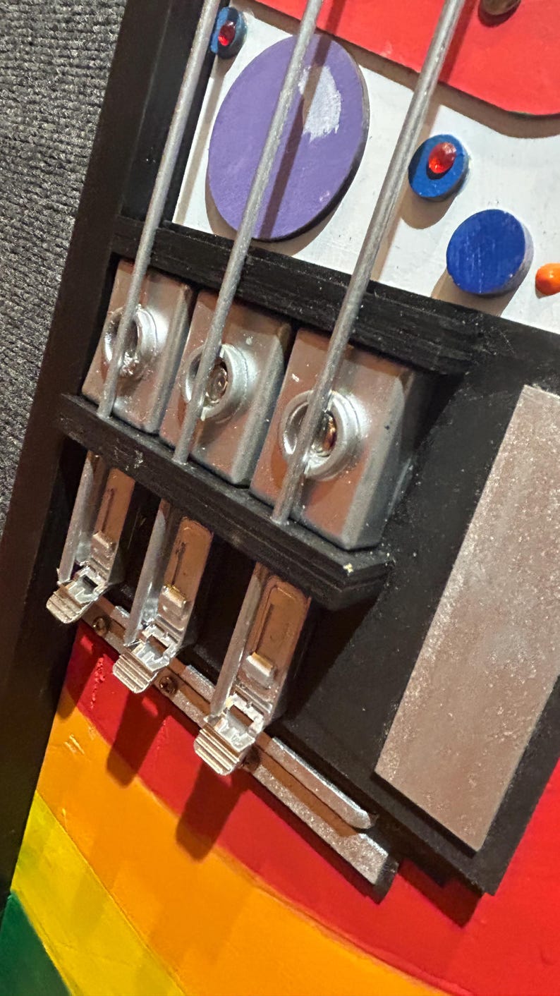 Może przedstawiać: Zbliżenie kolorowego, abstrakcyjnego dzieła sztuki przypominającego gitarę basową. Grafika przedstawia tęczowy korpus, srebrne struny i metalowy osprzęt. Elementy dekoracyjne obejmują fioletowe, niebieskie i czerwone koła.