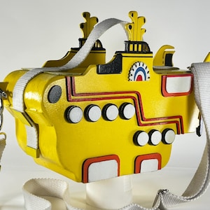 Può includere: Borsa gialla a forma di sottomarino con cinturino bianco. La borsa presenta dettagli rossi e neri, oblò bianchi e un emblema decorativo. La borsa è progettata per assomigliare all'iconico Yellow Submarine.