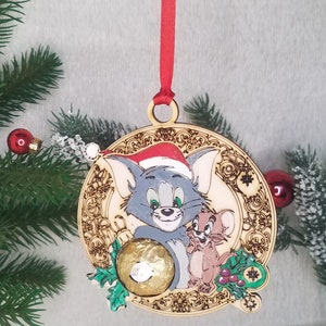 May include: A wooden Christmas ornament featuring Tom and Jerry. Tom is wearing a Santa hat and holding a gold ball. Jerry is standing next to Tom. The ornament is decorated with a green holly sprig and snowflakes.