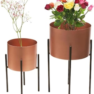 May include: Two copper plant pots with black metal stands. The larger pot has a bouquet of colourful roses. The smaller pot has a sprig of white flowers.