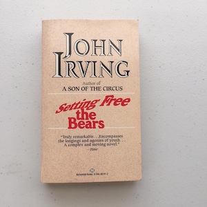 May include: A paperback book cover with a tan background. The title "Setting Free the Bears" is in red and white text. The author, John Irving, is in black text. The book is by the author of "A Son of the Circus".