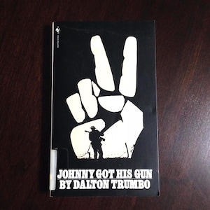 May include: A black and white book cover with a peace sign and a silhouette of a person holding a gun. The title of the book is "Johnny Got His Gun" by Dalton Trumbo.