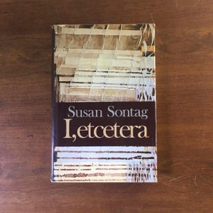 May include: A book titled "I, etcetera" by Susan Sontag. The cover features a watercolor-style abstract design in shades of brown, beige, and white.