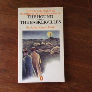 May include: A vintage paperback book cover featuring an illustration of two men walking on a desolate moor under a cloudy sky. The book title is "The Hound of the Baskervilles" by Sir Arthur Conan Doyle.