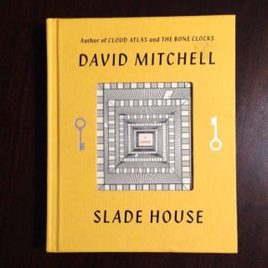 May include: A hardcover book titled "Slade House" by David Mitchell. The book has a bright yellow cover with the author's name and title in black. The cover features a square cutout revealing a black and white geometric design. Two key illustrations are on the cover.