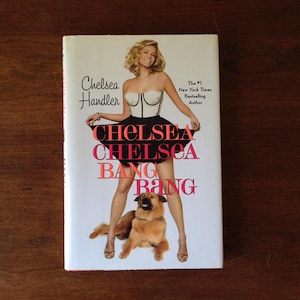 May include: A book cover with a woman in a white corset and black skirt, holding the skirt up. The title of the book is "Chelsea Chelsea Bang Bang" in large, pink letters. The author's name, "Chelsea Handler", is printed in black letters above the title. The book cover also includes the text "The #1 New York Times Bestselling Author". A brown dog is sitting in front of the woman.