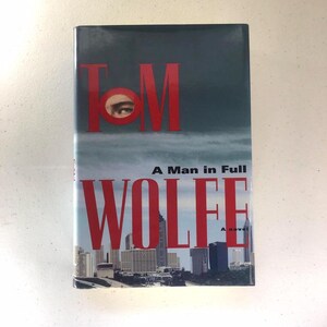 May include: A book cover with the title "A Man in Full" by Tom Wolfe. The cover features a red and white cityscape with a close-up of an eye in the "O" of the author's name.
