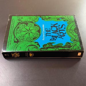 Może przedstawiać: Książka zatytułowana "The Black Arts" autorstwa Richarda Cavendisha. Okładka ma zielono-niebieski wz&oacute;r z geometrycznymi kształtami i tekstem. Książka jest w czarnym etui.