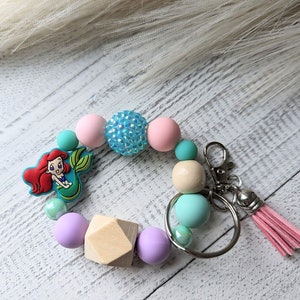 Puede incluir: Un llavero de cuentas con un dije de sirena, cuentas pastel en rosa, azul y lavanda, y una cuenta geométrica de madera. El llavero incluye un cierre plateado y una borla rosa.