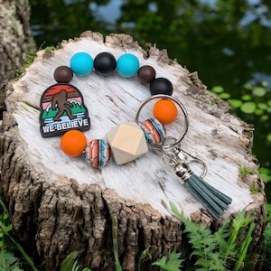 May include: A colorful beaded keychain with a wooden bead, a metal key ring, and a charm with a cartoon Bigfoot and the text "WE BELIEVE".