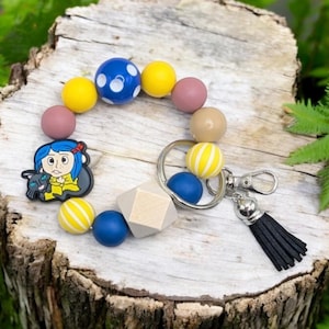 May include: A colorful beaded keychain with a charm of a girl with blue hair and a black cat. The keychain has yellow, blue, brown, and white beads. The charm is attached to a silver key ring with a black tassel.