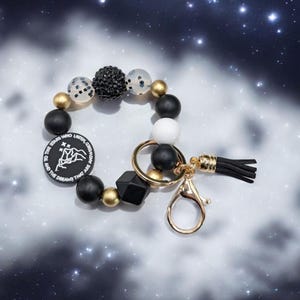 May include: A beaded keychain wristlet in black, white, and gold. The wristlet features a variety of beads, including round and geometric shapes, with a black tassel and a gold-colored clasp. A black circular charm has the text "TO THE STARS WHO LISTEN, AND THE DREAMS THAT ARE ANSWERED."