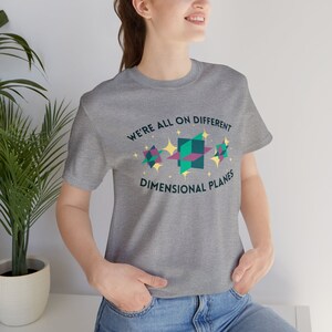 May include: A gray t-shirt with the text "WE'RE ALL ON DIFFERENT DIMENSIONAL PLANES" in a circular design. The design includes geometric shapes in teal, pink, and yellow with star accents. The shirt is made of a soft material.