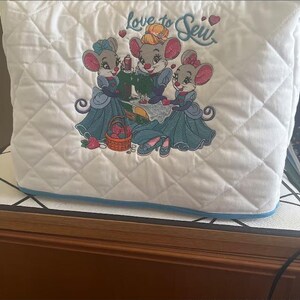 May include: White quilted sewing machine cover with a blue trim. The cover features an embroidered design of three mice dressed in blue dresses and the text "Love to Sew".