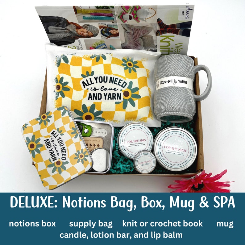 A deluxe knitting and crochet gift box is shown on a white background.  Inside the knitting gift box are a notions tin, mug, supply bag, candle, lotion bar, and lip balm.