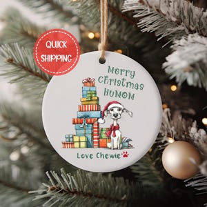 May include: White ceramic Christmas ornament with a cartoon dog wearing a Santa hat and a scarf, standing next to a stack of wrapped presents. The ornament reads "Merry Christmas Humom Love Chewie".