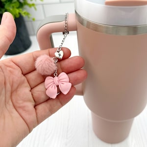 May include: A pink tumbler with a silver chain attached to the lid. The chain has a pink bow, a pink pom-pom, and a silver heart charm with the letter "R" on it.