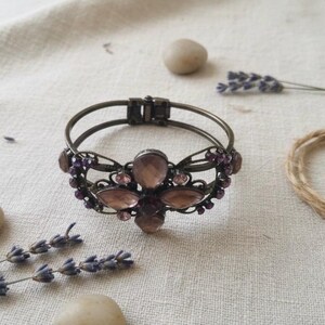 May include: A vintage-style bracelet with a bronze finish. The bracelet features a central floral design with large, oval-shaped, light purple gemstones and smaller purple accent stones. Lavender sprigs and small stones are arranged around the bracelet.