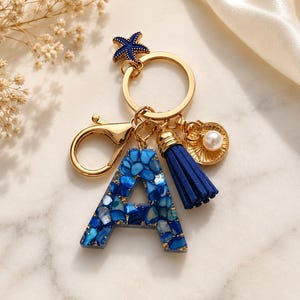 May include: A gold-toned keyring featuring a letter A charm filled with blue stone fragments and gold flakes. The keyring includes a dark blue tassel, a small gold shell charm with a white pearl, and a dark blue starfish charm attached to a ring.