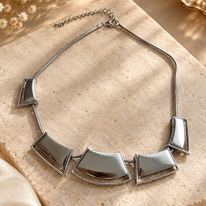 May include: A silver-toned necklace featuring a snake chain and five curved, polished metal plates. Each plate is framed by a twisted rope-style border. The necklace includes an adjustable chain with a lobster claw clasp closure.