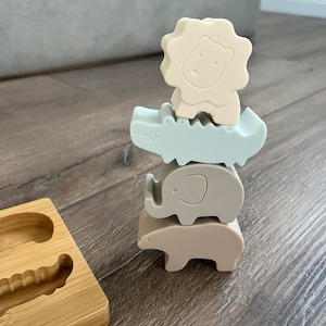 Puede incluir: Un conjunto de cuatro juguetes apilables de silicona de colores con forma de león, cocodrilo, elefante y oso. Los juguetes están apilados uno encima del otro sobre una superficie de madera.