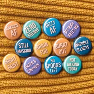May include: A set of 12 round button pins with various phrases related to feeling unwell, such as "Anxious AF", "Zero Energy", "Dizzy AF", "Might Faint", "Still Masking", "Brain Fog", "Burnt Out", "Dazed & Confused", "No Spoons Left", "My Body Hates Me", "Invisible Illness", and "Not Talking Today".