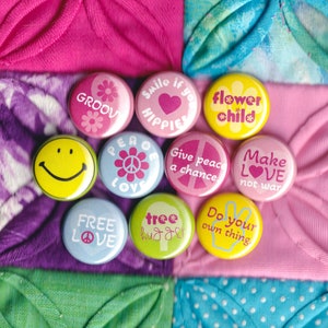 Könnte beinhalten: Neun bunte runde Knöpfe mit Friedens- und Liebesbotschaften. Die Knöpfe befinden sich auf einem Patchwork-Hintergrund aus rosa, blauem und grünem Stoff.