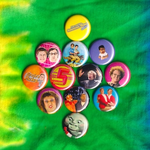 May include: A collection of 11 round button pins featuring various characters and logos from popular television shows. The pins are arranged in a circular pattern on a green and yellow tie-dye fabric background. The pins feature characters from shows like "Tim and Eric Awesome Show, Great Job!" and "The Fifth Element".