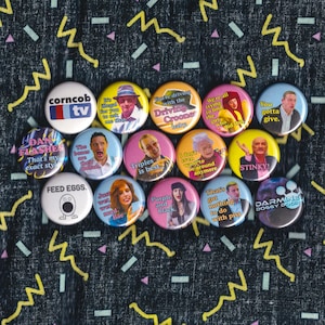 May include: A collection of colorful round pinback buttons with various images and text. The buttons feature phrases like "FEED EGGS" and "STINKY!" along with images of people. The background is a dark fabric with a geometric pattern.