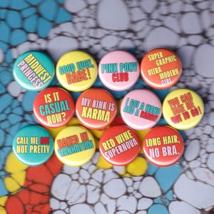 Może przedstawiać: Kolekcja kolorowych, okrągłych guzików z różnymi frazami pogrubioną czcionką. Guziki są w odcieniach czerwieni, różu, żółci, zieleni i pomarańczy. Frazy obejmują "Midwest Princess", "Good Luck Babe!" i "Pink Pony Club".
