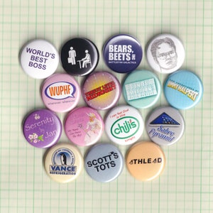 May include: A collection of 14 round button pins with various designs and text. The pins feature characters from popular TV shows and movies, including Serenity, Battlestar Galactica, The Office, and The Adventures of Jimmy Halpert. Other pins include the text "WUPHF", "Three Level Midnight", "I Can Feel God in This Chili's", "Vance Refrigeration", "Scott's Tots", "Sabre Pyramid", and "Athlead".