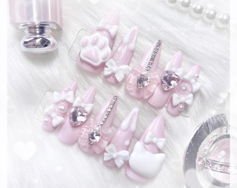 Peut inclure: Ensemble d'ongles artificiels roses avec diverses décorations. Certains ongles présentent des nœuds blancs, des strass et des pierres précieuses en forme de cœur. Un ongle a une empreinte de patte blanche et un autre une tête de chat blanche. Un flacon de vernis à ongles est visible.