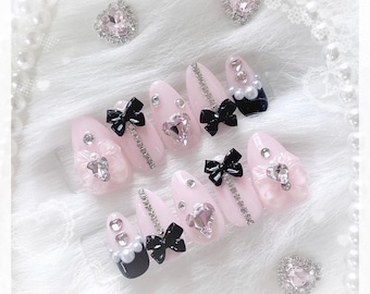 Peut inclure: Ensemble de faux ongles roses pâles, ornés de nœuds noirs, de strass et de gemmes en forme de cœur. Certains ongles présentent des perles et une base bleu marine foncé. Présentés sur une surface blanche et douce.