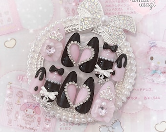 Peut inclure: Un ensemble d'ongles artificiels noirs et roses avec des strass et des motifs en forme de cœur. Les ongles sont présentés sur un plateau blanc perlé avec un nœud rose et une couronne.