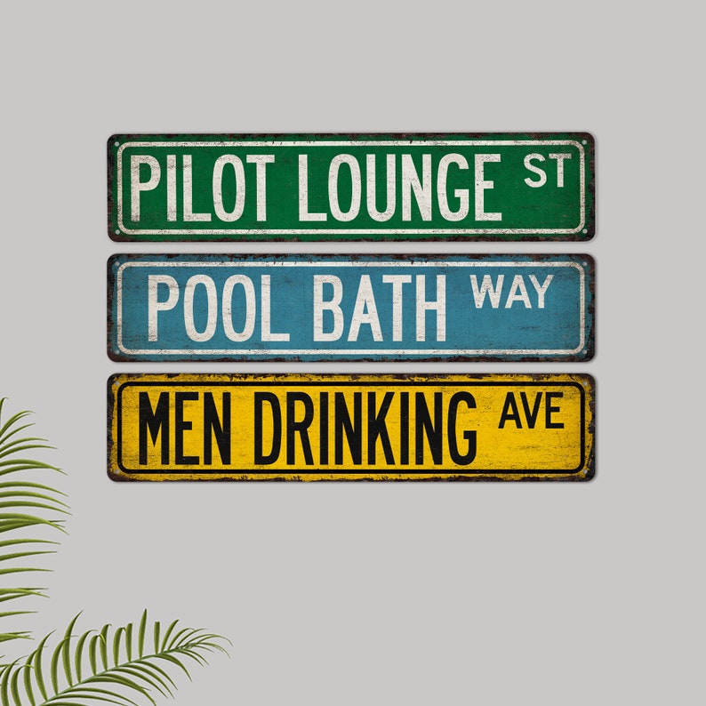 May include: Three vintage-style street signs with distressed paint. The top sign is green with white text that reads "Pilot Lounge St.". The middle sign is blue with white text that reads "Pool Bath Way". The bottom sign is yellow with black text that reads "Men Drinking Ave."