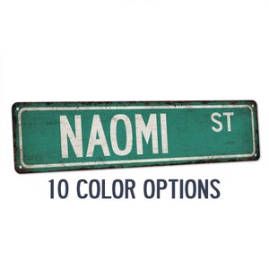 May include: A green metal street sign with the text "NAOMI ST" in white lettering. The sign has a distressed, rusty look. 10 color options are available.