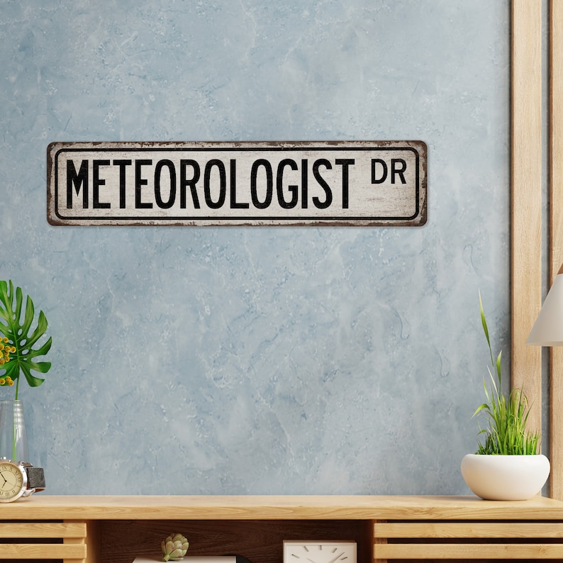 May include: A white metal sign with black text that reads "METEOROLOGIST DR". The sign has a distressed, vintage look.
