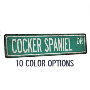 May include: Rustic metal sign with the text "Cocker Spaniel Dr." in white lettering on a green background. The sign has a distressed look and is perfect for a dog lover's home.