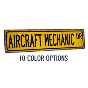 May include: A yellow and brown metal sign with the text "AIRCRAFT MECHANIC DR" in black letters.