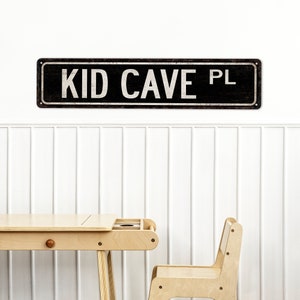 May include: A black and white sign with the text "KID CAVE PL" on a white wall. A light wood desk and chair are in the foreground.